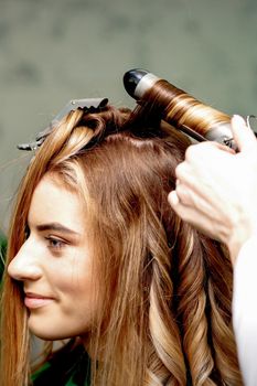 Professional hairdresser makes curls with a curling iron for a young woman with long red hair in a beauty salon