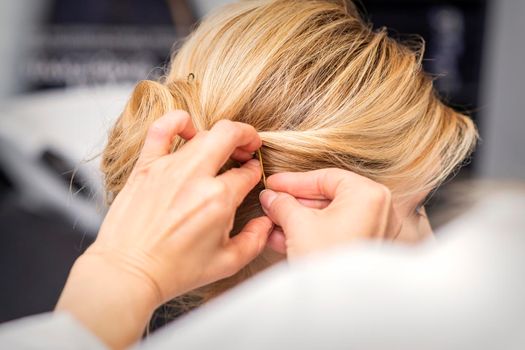 Closeup back view of a hairdresser stabs blonde hair with a hairpin for a young woman in a hair salon