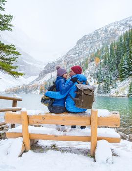 Lake Agnes by Lake Louise Banff national park is a lake in the Canadian Rocky Mountains. A young couple of men and women sitting on a bench by the lake during a cold day in Autumn in Canada with snow