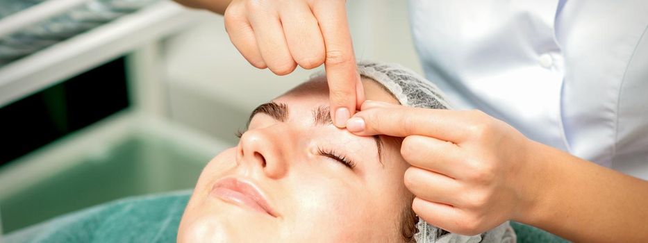 Beautician's hands make massage on eyebrow for a woman while a facial massage in cosmetology clinic center