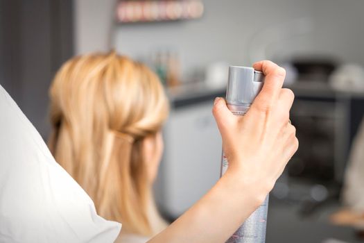 Closeup of hair spray in the hand of hairstylist while fixing blonde hair of the young woman in a beauty salon