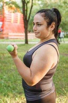 A charming brunette woman plus-size body positive practices sports in nature. Exercises with dumbbells.