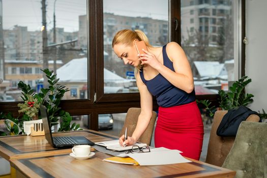 Young businesswoman talks on the phone writing notes standing in office or cafe