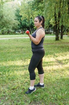 A charming brunette woman plus-size body positive practices sports in nature. Exercises with dumbbells.