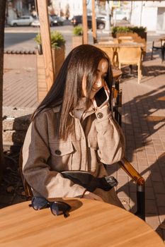A stylish brunette woman is sitting at a table in an outdoor cafe with coffee and a phone. The girl is talking on the phone. Autumn walk.