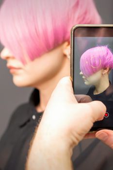 The hairdresser takes pictures of the short pink hairstyle of a young woman on the smartphone