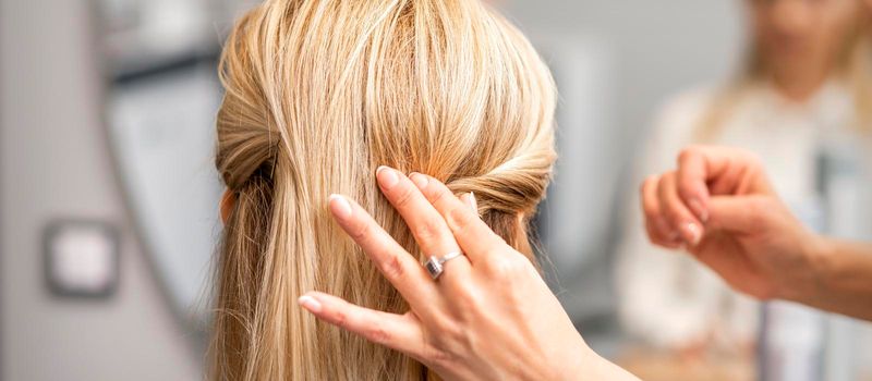Back view of the female hand of hairdresser models a hairstyle of a young blonde woman in a hair salon