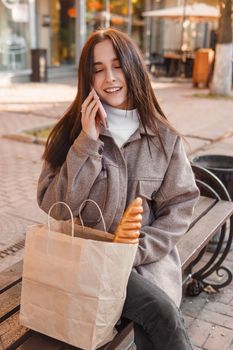 A stylish brunette woman walks around the autumn city. The brunette is sitting on a bench outside with a glass of coffee and a phone, holding a craft bag with a baguette.