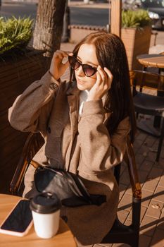 A stylish brunette woman in sunglasses is sitting at a table in an outdoor cafe with coffee and a phone. The girl is talking on the phone. Autumn walk.