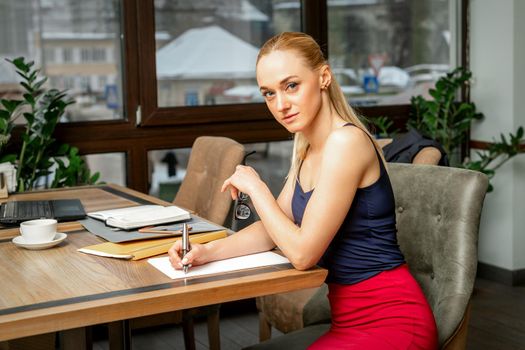 Beautiful young businesswoman is writing documents looking at camera sitting in a cafe