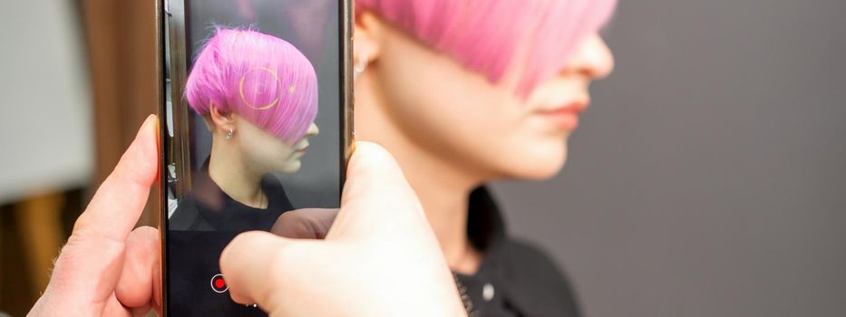 The hairdresser takes pictures of the short pink hairstyle of a young woman on the smartphone