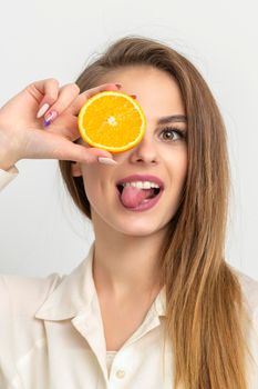 Portrait of a cheerful caucasian young woman covering eye with an orange slice and stick out tongue wears white shirt against a white background