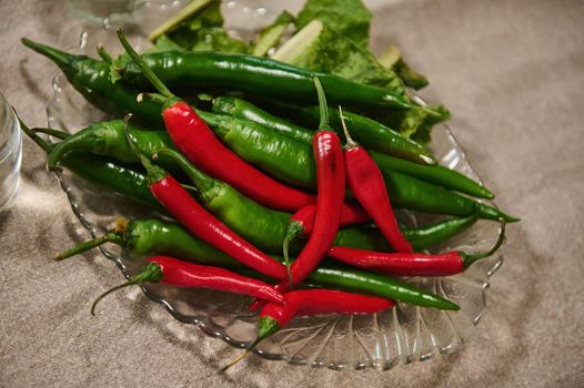 View from above of fresh raw red and green chili peppers in a plate on kitchen table with linen tablecloth. Raw vegan. Spicy ingredient. Still life