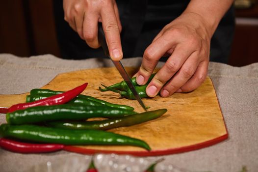 Details: Hands of chef or housewife using kitchen knife cutting red and green chili pepper on a chopping board. Slicing food, preparing spicy ingredients for canning or pickling. Raw vegan vegetables