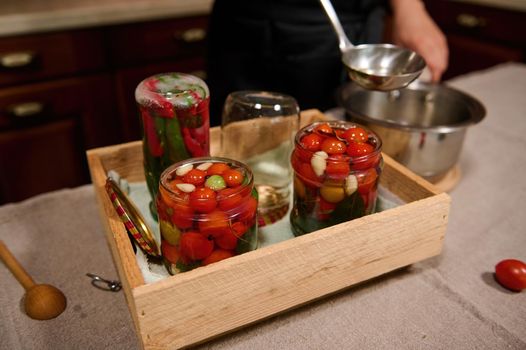 Focus on sterilized cans for canning filled with cherry tomatoes, on the background of a chef using soup ladle, pours boiling brine from a saucepan into jars, preparing fermented delicacies for winter