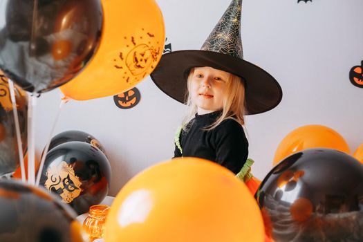 Children's Halloween - a girl in a witch hat and a carnival costume with airy orange and black balloons at home. Ready to celebrate Halloween.