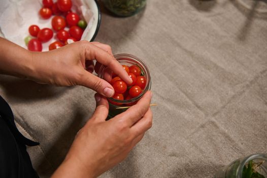 View from above of women's hands putting ripe organic homegrown cherry tomatoes in a sterilized jar for canning. Preparing fermented vegetables, according to traditional family recipe at home kitchen