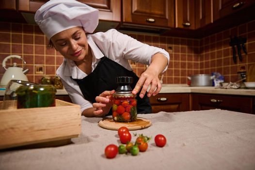 Beautiful Hispanic housewife in white chef's cap and black apron, using seamer, closes lid of jar with ripe and juicy tomatoes while canning according to traditional family recipe in home kitchen