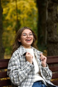 European elegant young woman in a stylish trench coat in a white sweater and blue jeans resting on the bench. warm autumn day. brown wooden bench