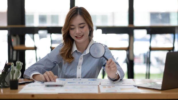 Young Asian businesswoman working on a laptop computer with a calculator and examining a contract meticulously with a magnifying glass at office.