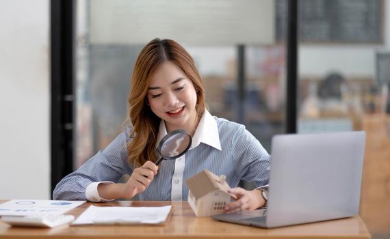 Close up of businesswomen holding magnifying glass finding a wooden shape of a house, realtor, agent, insurance, developer, planning and investment real estate business concept,.