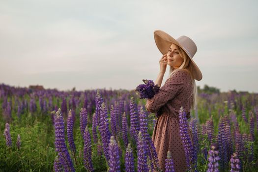 A beautiful woman in a straw hat walks in a field with purple flowers. A walk in nature in the lupin field.