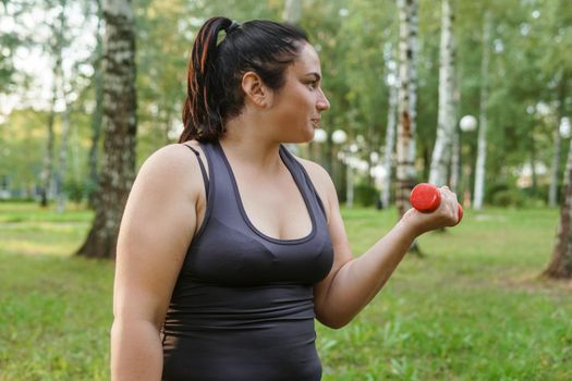 A charming brunette woman plus-size body positive practices sports in nature. Exercises with dumbbells.