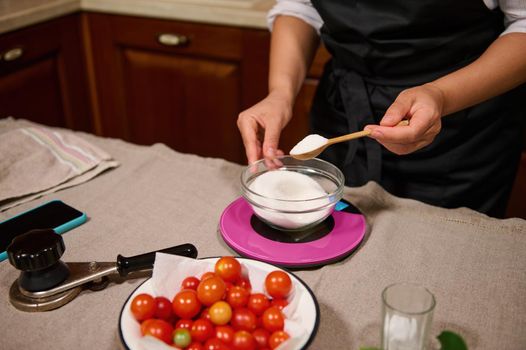 View from above of the hand of a housewife in black chef's apron holding wooden teaspoon while weighing sugar on kitchen scale, preparing marinade and homemade pickles for the winter. Canning food