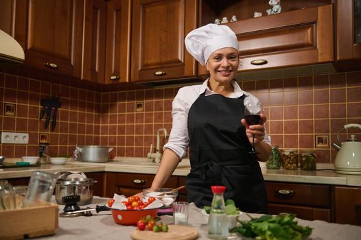 Beautiful Hispanic housewife woman in white cap and back apron, holding a glass of red wine and smiling cutely at camera, standing in a cozy wooden home kitchen with ingredients on a kitchen island