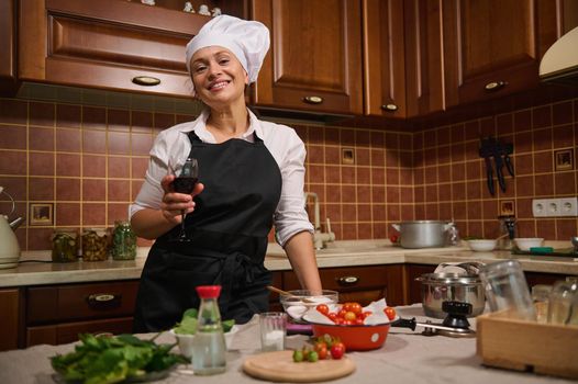 Charming middle-aged Hispanic woman, housewife in black apron and white chef's cap, sipping a glass of red wine, smiles at camera while cooking at home kitchen, preparing canned food for the winter