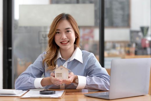 Miniature house in the hands of an Asian woman real estate agent home loan working at the office. Looking at the camera..