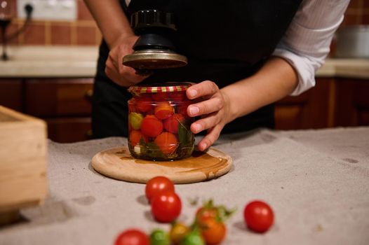 Cropped view of housewife using a special key to close the lids on jars of cherry tomatoes during winter preservation in the home kitchen. Traditional family recipe. Mediterranean culture and cuisine