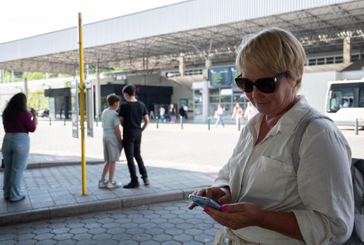 a middle-aged caucasian woman is waiting for a bus at a bus stop and writing a message on her mobile phone. High quality photo