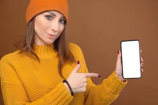 Smiling caucasian young lady pointing with finger at smartphone in hand, standing against orange studio background.