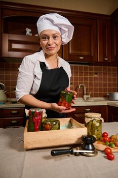 Pleasant multiethnic woman, housewife in white chef's hat and black apron, holding a jar of fermented cherry tomatoes upside down, smiling cutely at camera while standing at kitchen table. Canning