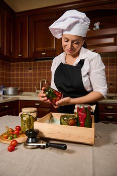 Cute woman, housewife in white chef's cap and black kitchen apron, stacking jars with homemade canned fermented and marinated vegetables in a wooden crate. Pickling according family traditional recipe