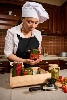 Beautiful woman, pleasant housewife in a white chef's hat and a black apron, stands at the kitchen table and puts a jar of fermented cherry tomatoes upside down on a wooden box with canned vegetables