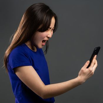 Angry asian woman screaming down, swears with somebody. Using mobile phone. Stressed and depressed girl on grey background.