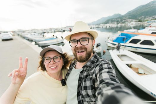 Young couple taking a self portrait laughing as they pose close together for camera on their smartphone outdoors in summer port with boats and yachts.