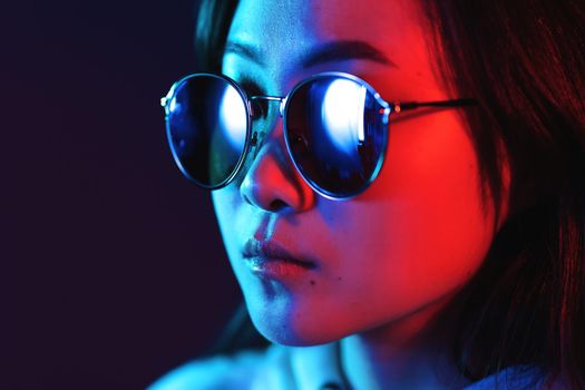 Young pretty girl with asian appearance in neon pink and blue lights at night. Serious trendy hipster teenager in glasses.