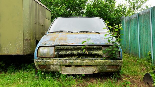 An old faulty car in an abandoned farm. Blue abandoned broken-down car against a background of blue fence and grass.