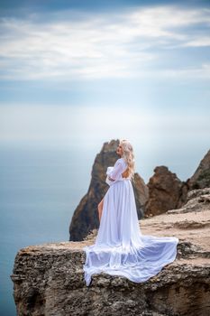 Blonde with long hair on a sunny seashore in a white flowing dress, rear view, silk fabric waving in the wind. Against the backdrop of the blue sky and mountains on the seashore