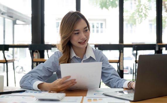 Young Asian businesswoman working on a laptop computer with a calculator in a bright modern office. Planning to analyze charts, business plans, investments, financial or accounting concepts..