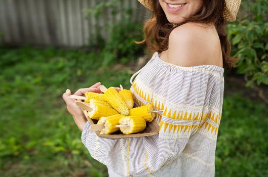 The girl is smiling in a straw hat and holding a plate with boiled corn in her hands. The concept of outdoor recreation, barbecue