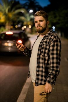 Man waits taxi by using transportation app on night street. Technologies and city