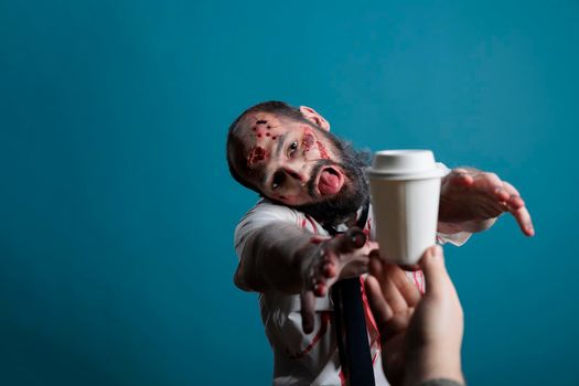 Aggressive halloween corpse wishing to drink coffee cup in studio, going after beverage and wanting to eat carbon. Spooky undead scary devil chasing hand with drink, acting dangerous and creepy.