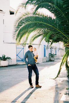 Dad with a baby in his arms stands near huge palm leaves in the courtyard of the house. High quality photo