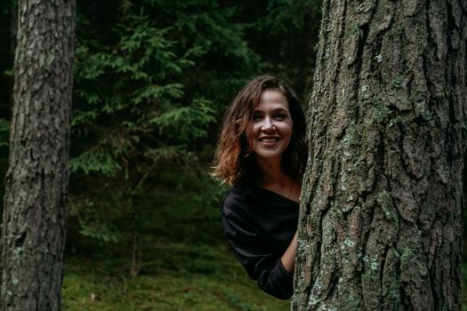 A young woman in a coniferous forest looks out from behind a tree. Happy young woman looking at the camera and smiling