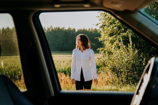 A beautiful young woman in a white shirt is resting outside the city. Outdoors near the forest and field. Photo through the car window. Car trip