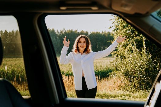 A beautiful young woman in a white shirt is resting outside the city. She is happy and laughs. Photo through the car window. Car trip